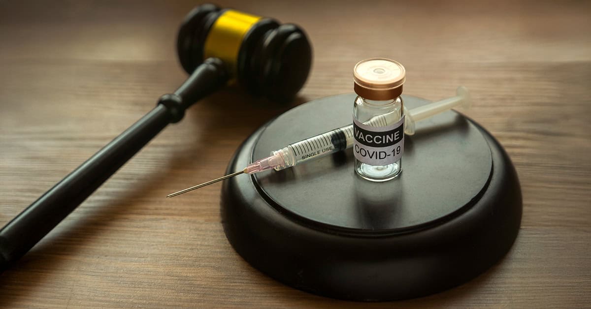Vaccine COVID-19 vial and syringe on a black platform, with a gavel in the background, symbolizing legal and medical aspects of the COVID-19 pandemic response.