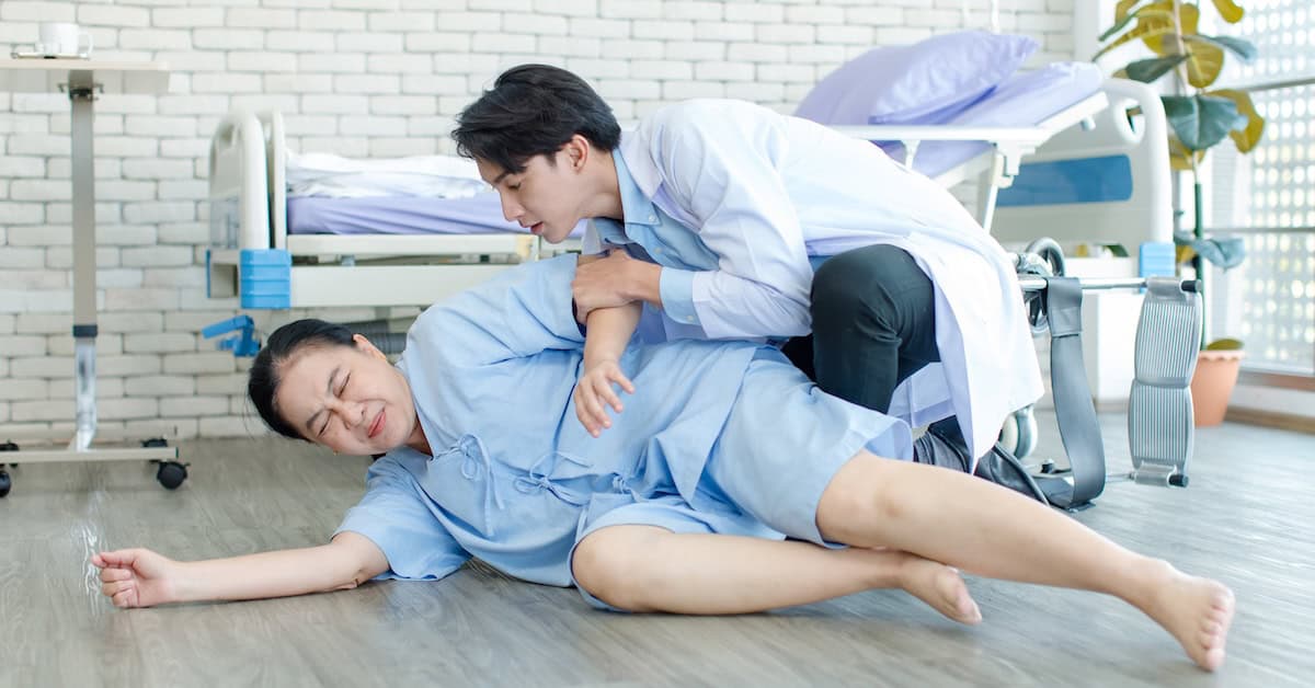 Young male doctor assisting late middle-aged woman experiencing a medical emergency on hospital floor, emphasizing urgent care and emergency response.