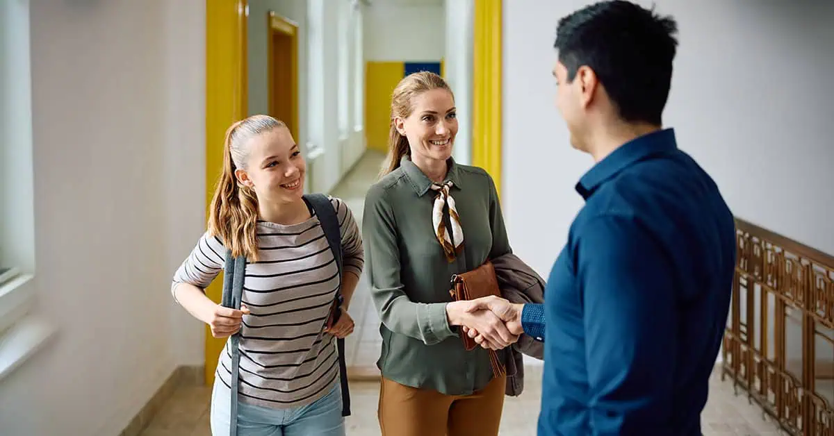 Happy mother of teenage girl shaking hands with high school principal in hallway